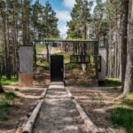 Mirrored cabin facade reflecting Scots pines, with a living grass roof, a black door, and a gravel path edged with logs