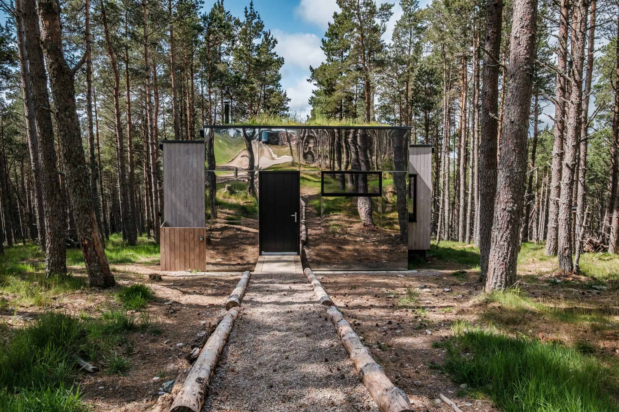 Mirrored cabin facade reflecting Scots pines, with a living grass roof, a black door, and a gravel path edged with logs
