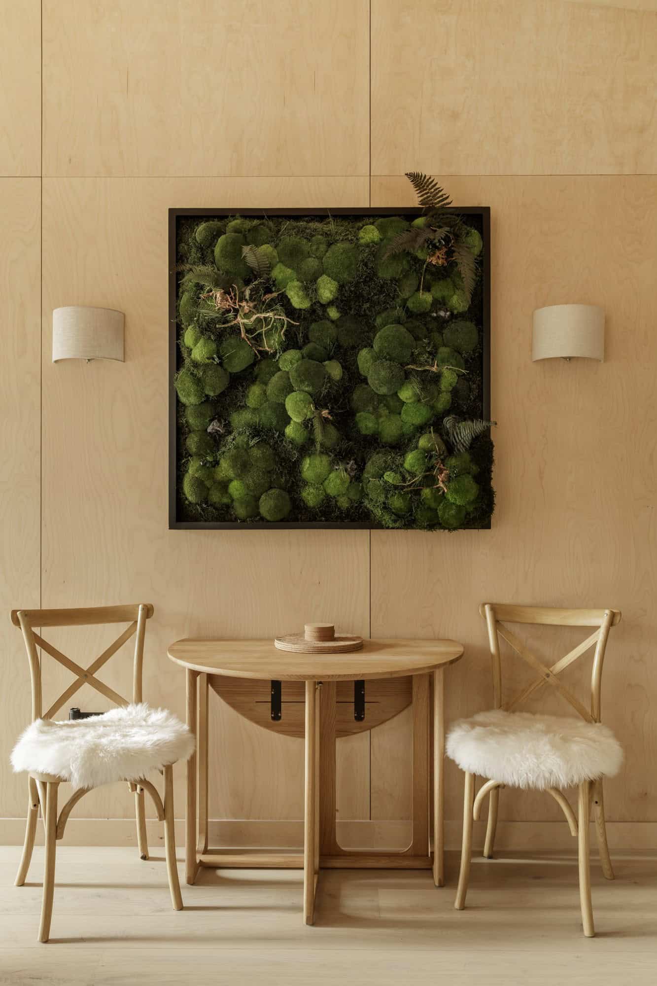 Light plywood cabin interior with a small oak table, two sheepskin chairs, wall sconces, and a framed moss artwork