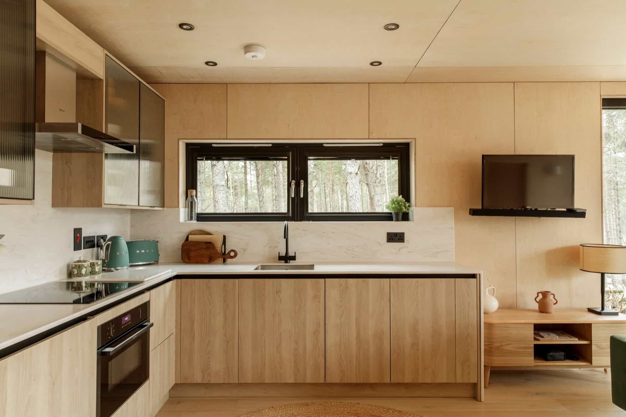 Light plywood kitchen with a white worktop, oak cabinets, a black tap, and a forest window above the sink