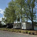 Three tiny houses on wheels behind a low rail fence under tall pines, gravel in front, a dark shed-roof house at left, a white gable with a porch in the middle, a pale grey house at right, and a lake with a fountain beyond at The Cottages at Pine Lake outside Huntsville