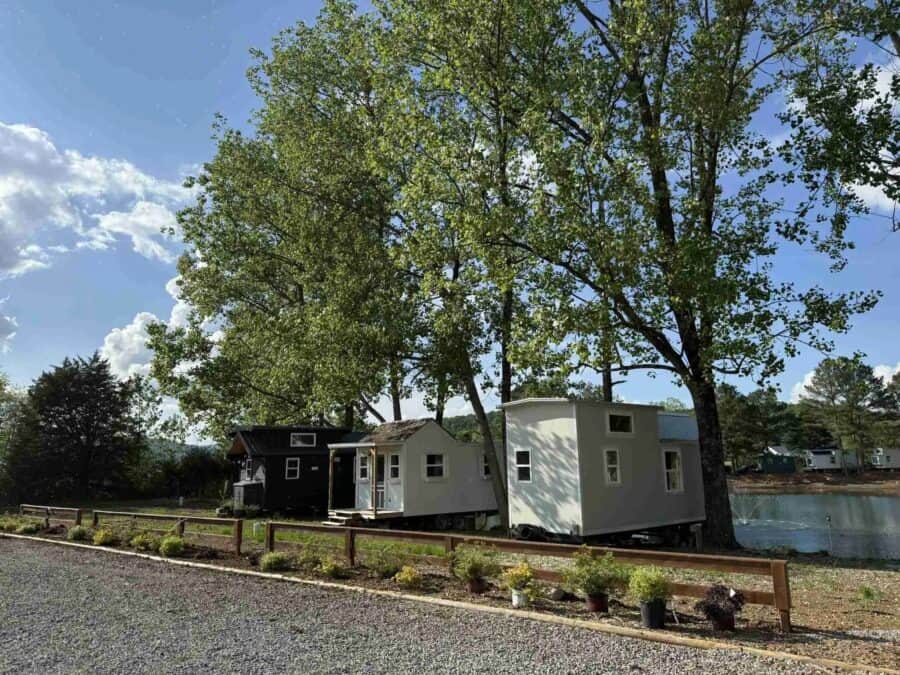 Three tiny houses on wheels behind a low rail fence under tall pines, gravel in front, a dark shed-roof house at left, a white gable with a porch in the middle, a pale grey house at right, and a lake with a fountain beyond at The Cottages at Pine Lake outside Huntsville