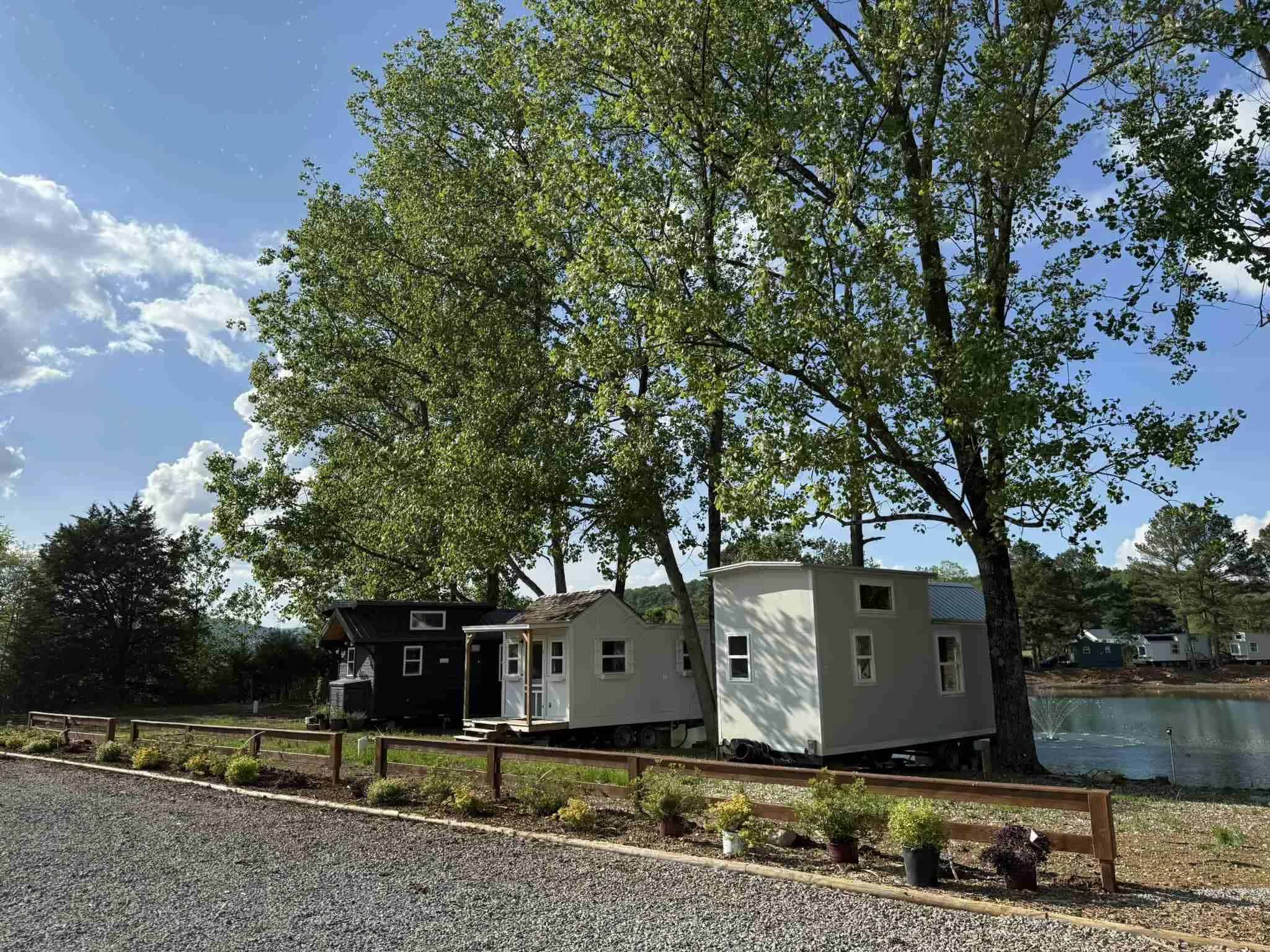 Three tiny houses on wheels behind a low rail fence under tall pines, gravel in front, a dark shed-roof house at left, a white gable with a porch in the middle, a pale grey house at right, and a lake with a fountain beyond at The Cottages at Pine Lake outside Huntsville
