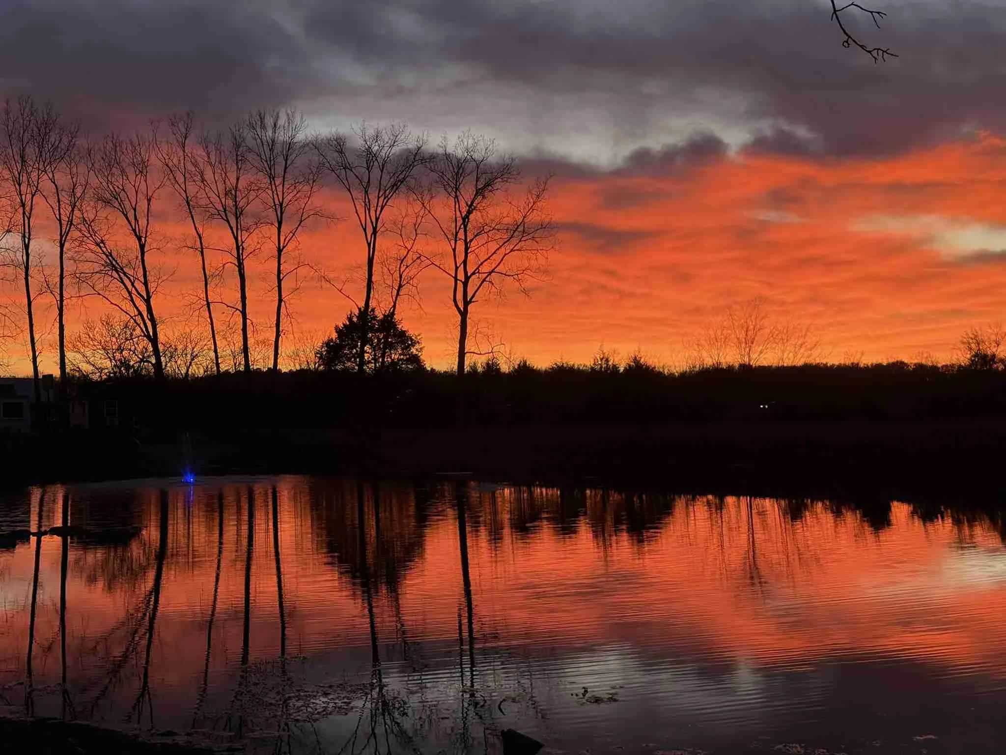 Sunset over the private lake at The Cottages at Pine Lake, orange sky mirrored in still water, a fountain lit blue at left, silhouetted trees and the corner of a house