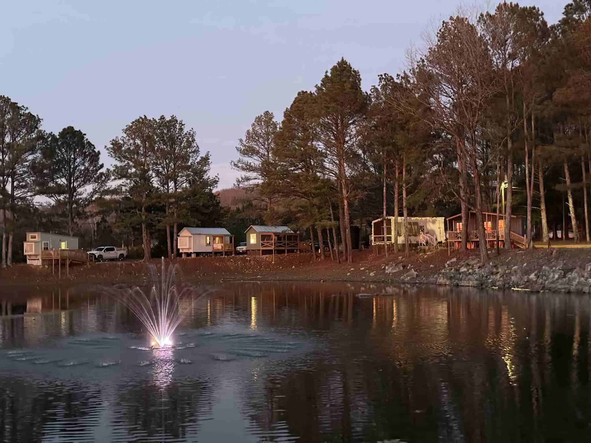 Twilight view of tiny houses along a rocky lake shore, windows lit, porches facing the water, a fountain on in the lake, and tall pines behind the row at The Cottages at Pine Lake
