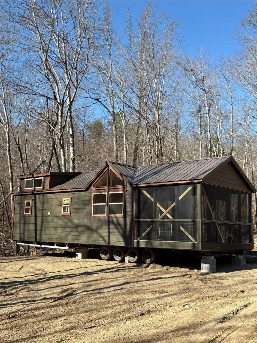 Cherokee cottage with dark green siding, a bronze metal roof, twin front gables, and a screened porch, set among bare trees at Cottages at Timber Crest in Bean Station, Tennessee
