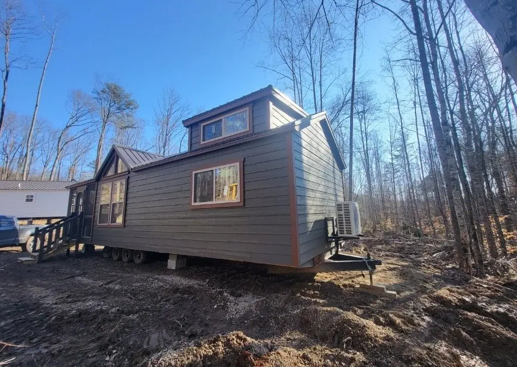 Side view of the Cherokee cottage showing its long green-sided body, dormer, and gabled porch end on a cleared wooded lot at Cottages at Timber Crest