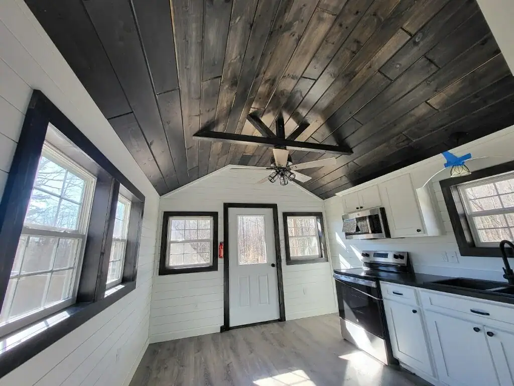 Woodland cottage interior with white shiplap walls, a dark plank ceiling with exposed trusses, black-trimmed windows, and a white kitchen