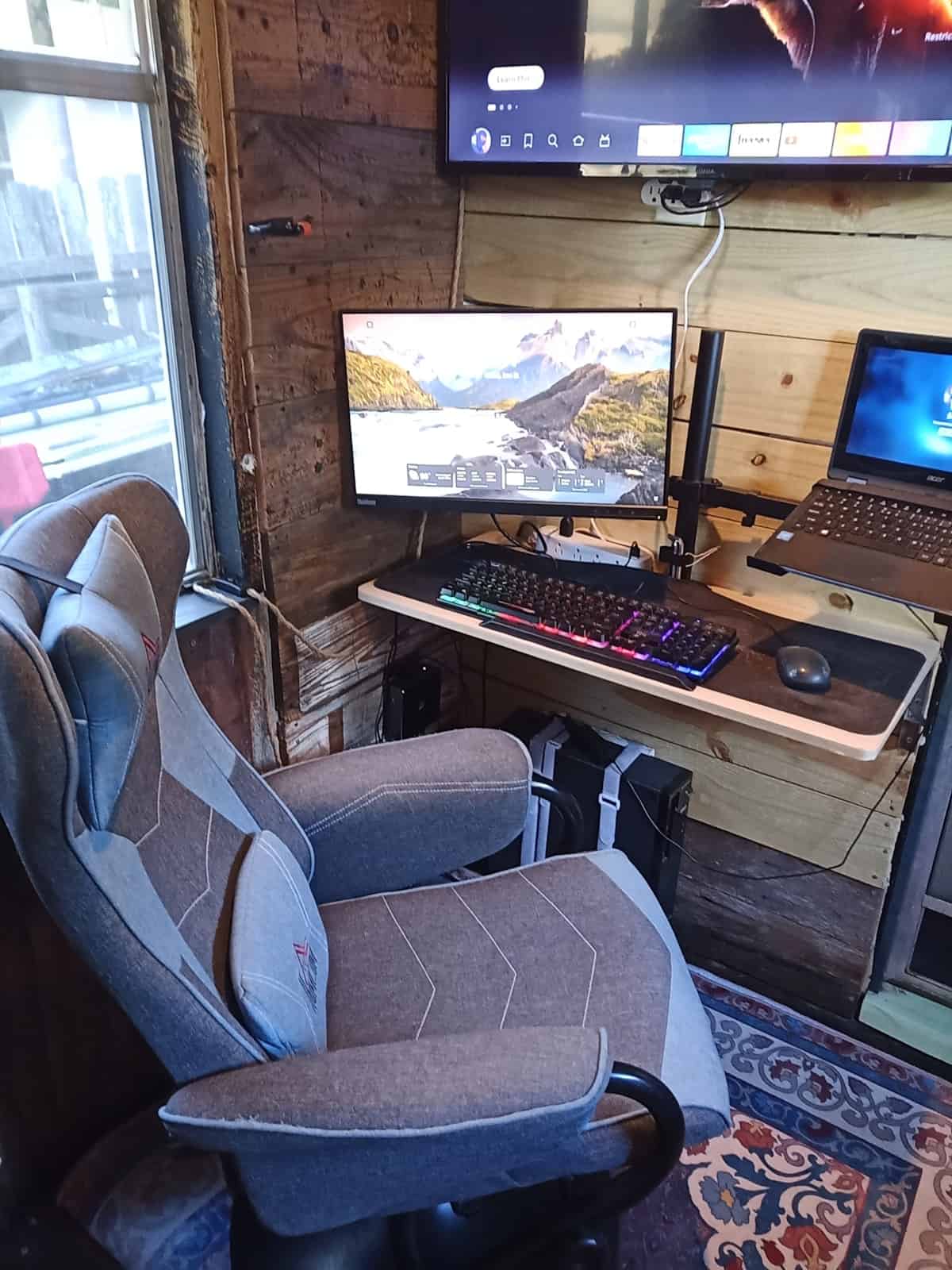 Office nook with a wood desk, monitor, laptop, high-back chair, wall TV, and a side window