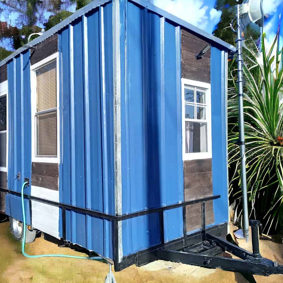 Hitch end of the blue corrugated tiny house showing the trailer tongue, a small front window, and wood trim