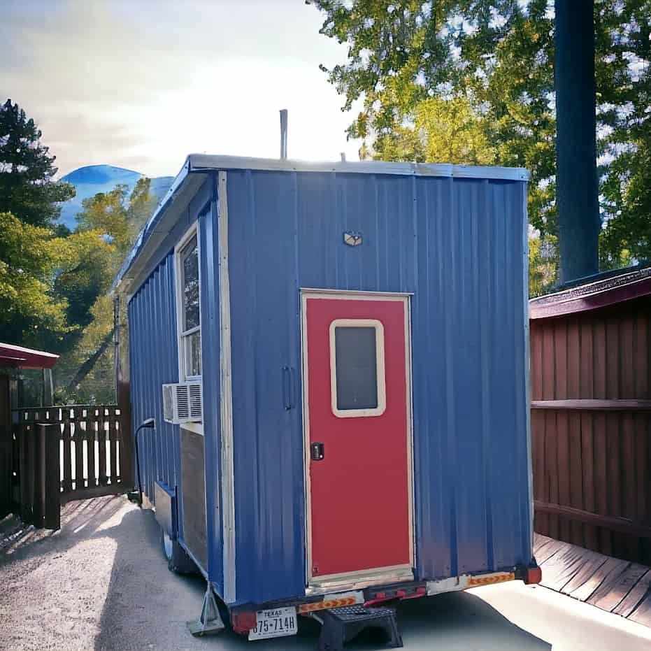 End view of the blue metal tiny house on a trailer, red door, window air conditioner, and Texas plate