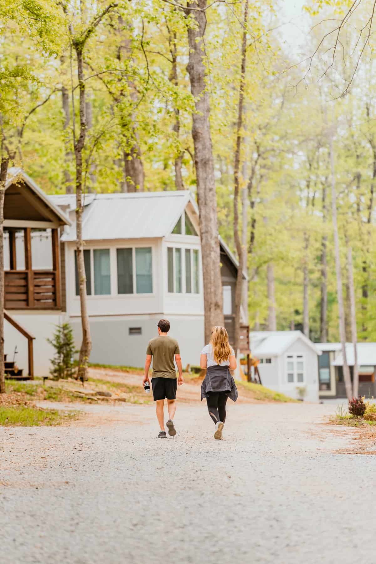 A couple walks down a gravel lane past gray and white park-model cottages under spring trees