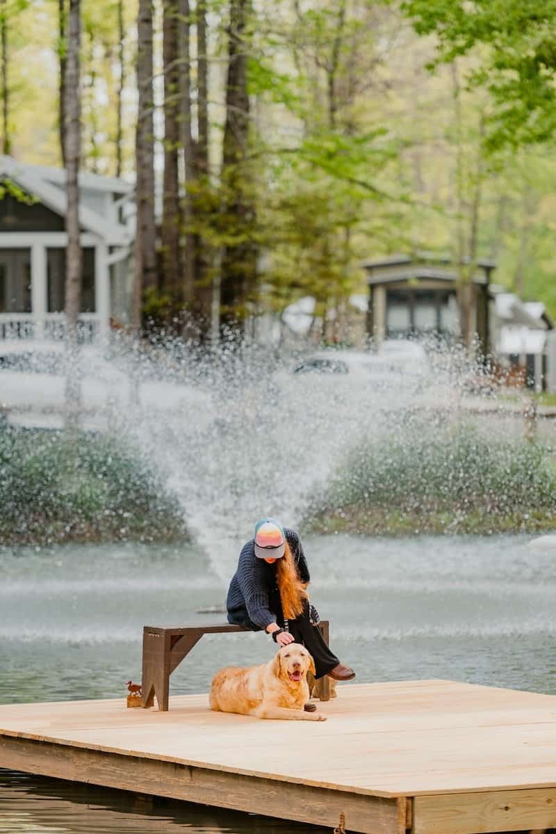 A resident sits on a dock with a golden retriever in front of a pond fountain