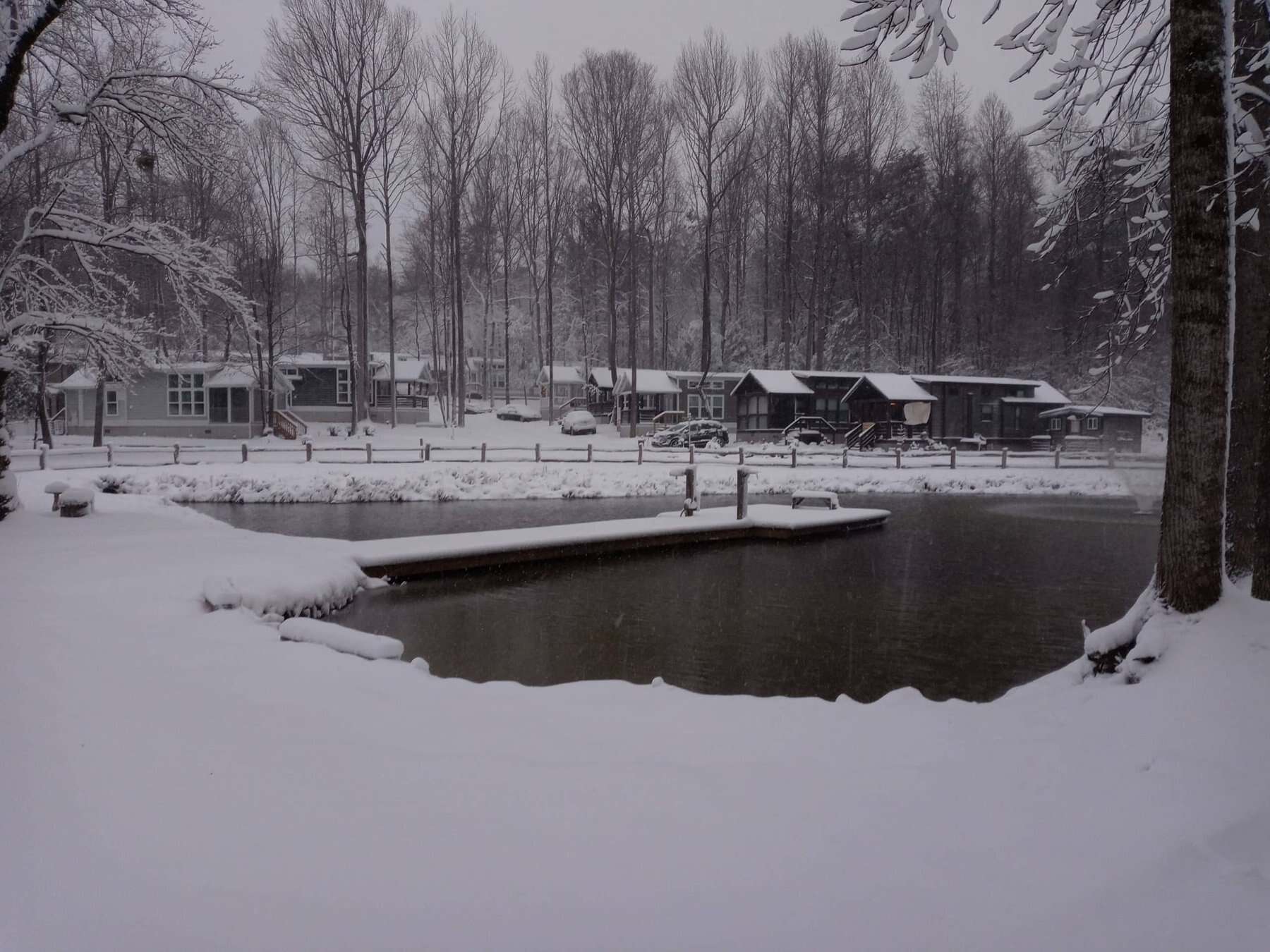 Snow-covered cottages and trees around a dark pond with a dock on a winter morning