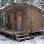 Dave Whipple's finished 10x16 off-grid cabin in snowy woods with porch chairs and glowing windows