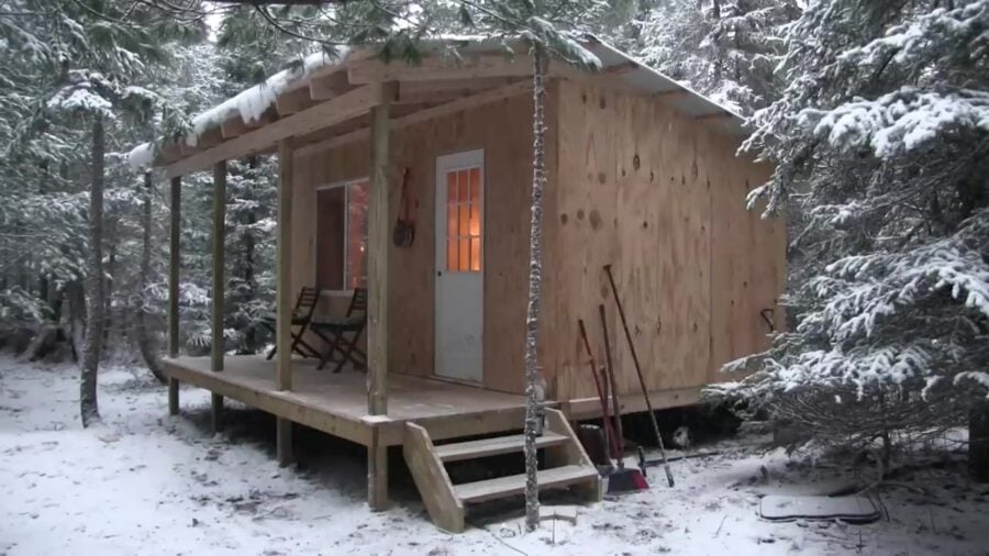 Dave Whipple's finished 10x16 off-grid cabin in snowy woods with porch chairs and glowing windows