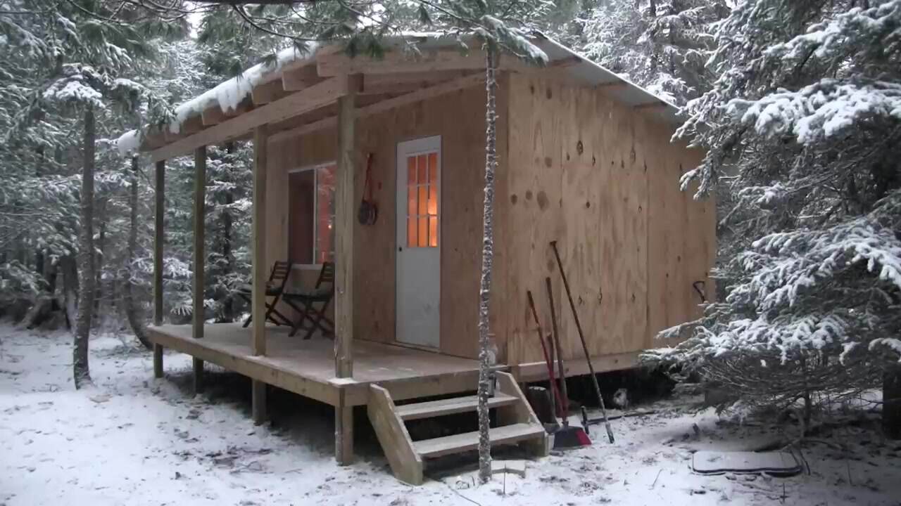 Dave Whipple's finished 10x16 off-grid cabin in snowy woods with porch chairs and glowing windows
