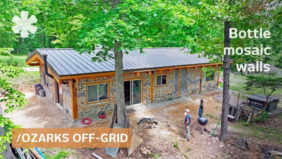 Cordwood and bottle-mosaic facade under a dark metal roof in a forest clearing, sliding door, dog and two people at a grill