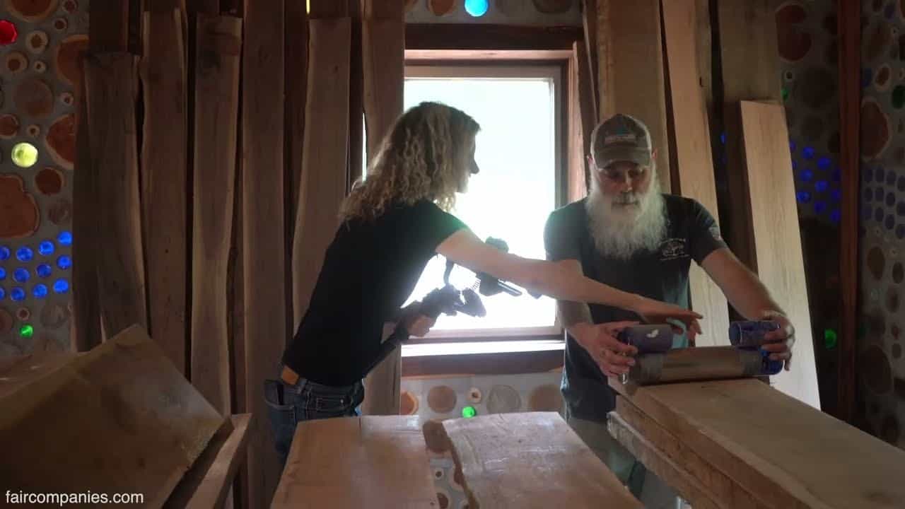 Unfinished interior with cordwood and colored bottle ends in mortar walls, live-edge slabs, Kirsten filming, Jason at a workbench