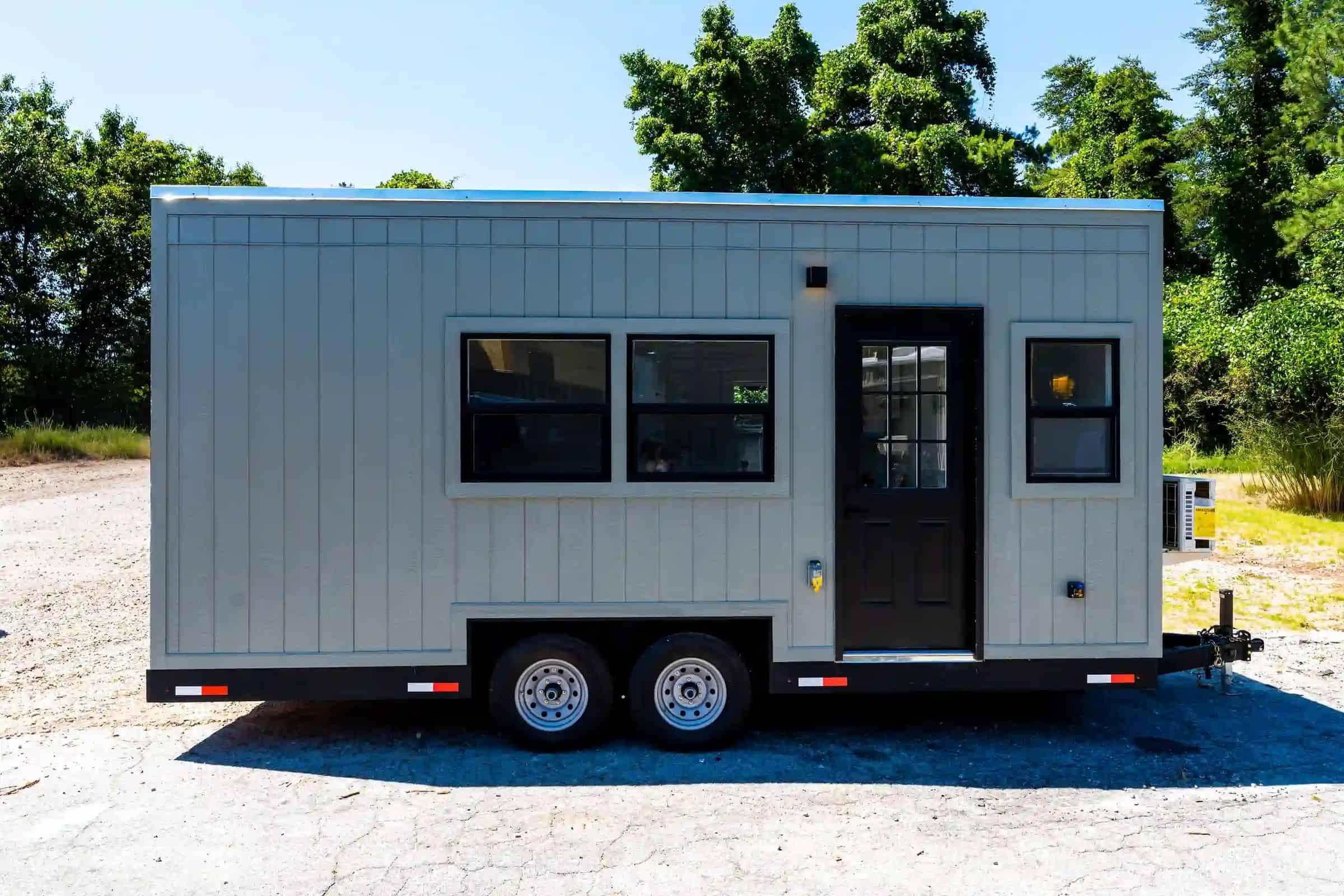 Side profile of a Fairfax-family THOW: grey vertical siding, black-framed windows, black door, dual-axle, hitch and mini-split visible