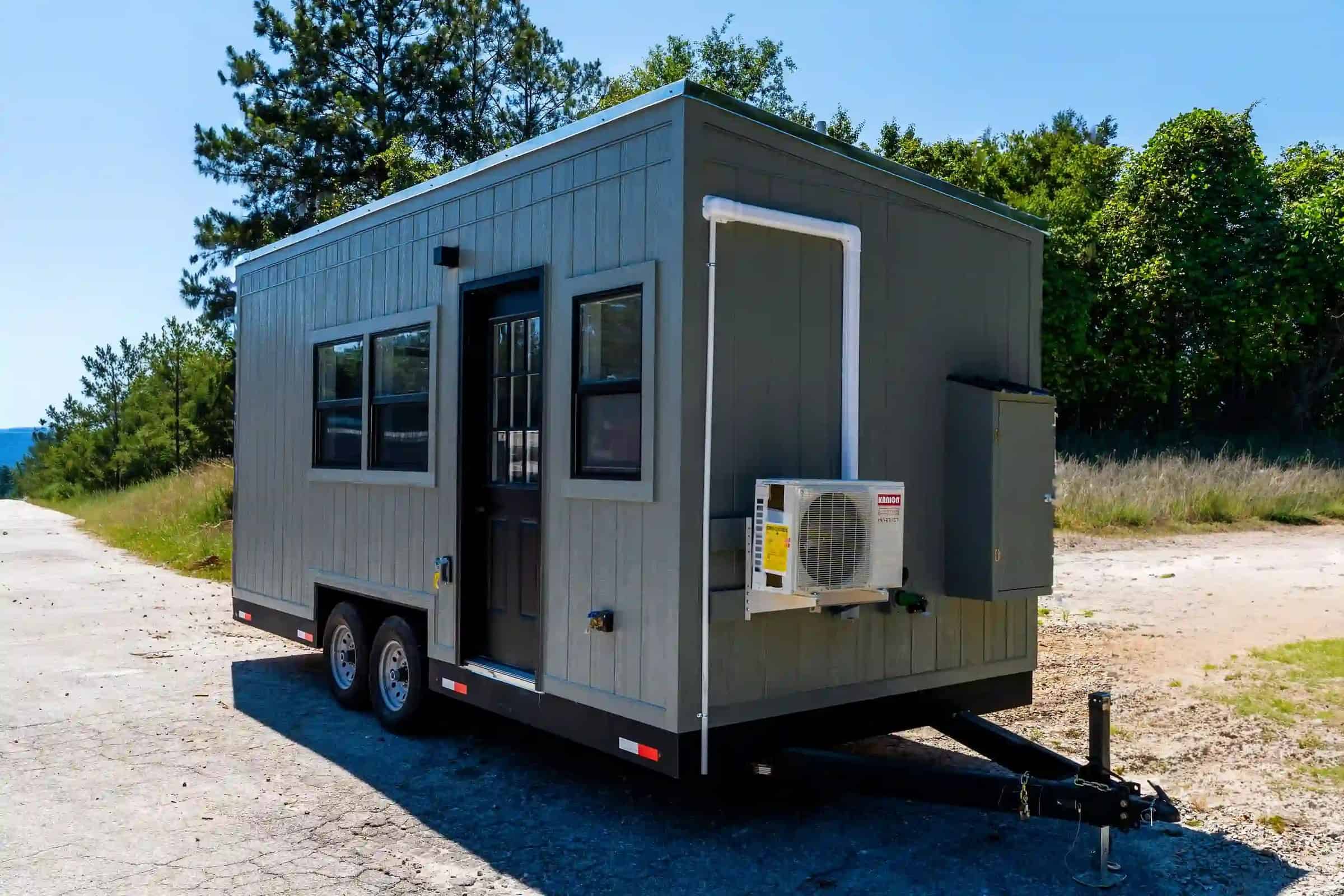 Three-quarter exterior of a Fairfax-family build: grey siding, black windows, dual-axle on gravel, white mini-split on hitch wall