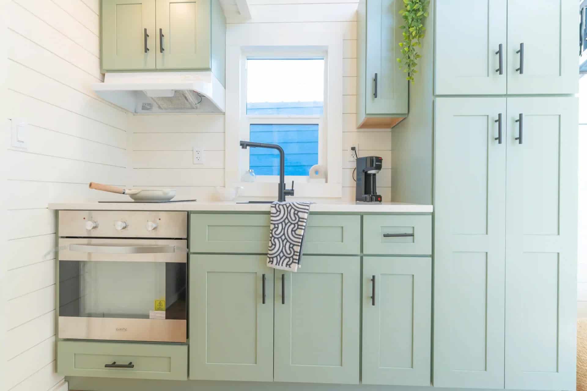 Genesis gallery kitchen: mint-green shaker cabinets, wall oven, two-burner cooktop, range hood, black faucet at a window, tall pantry