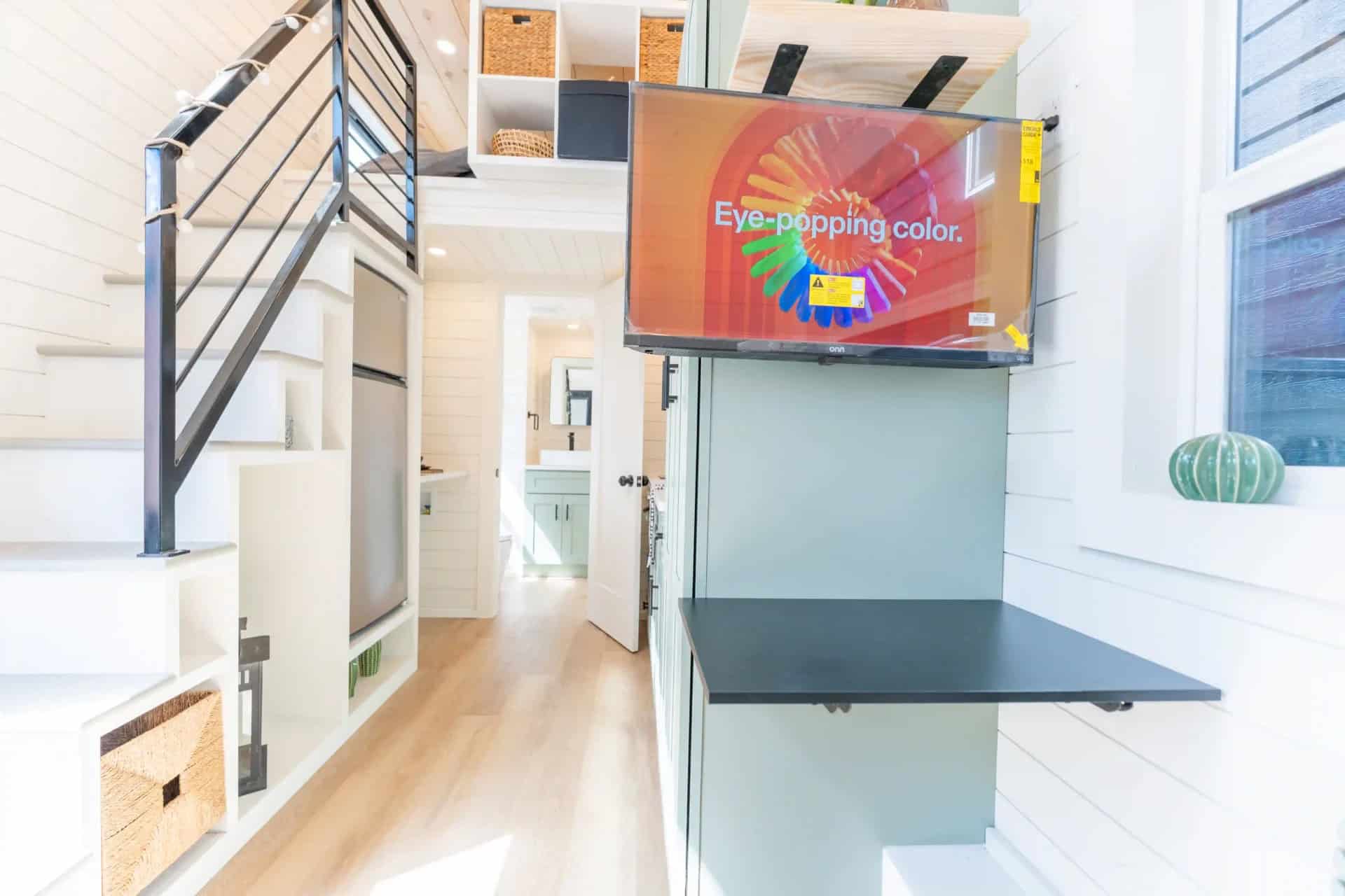 Genesis gallery interior: wall-mounted TV above a black fold-down desk on the mint cabinet wall, stairs and bath hallway at left
