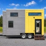Dragon Genesis 16 listing lead: grey horizontal siding and yellow board-and-batten THOW on a dual-axle trailer, black glass door, wildflower field behind