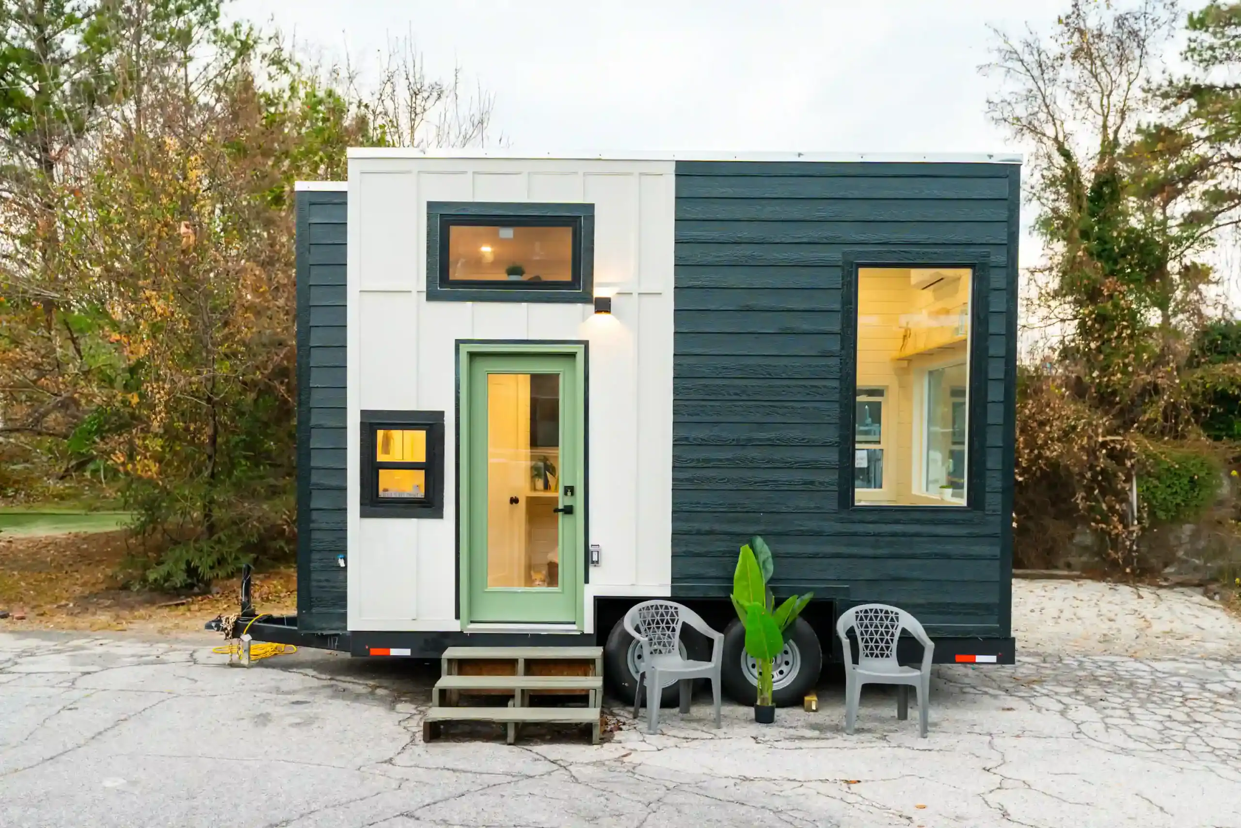 Sora exterior with a sage-green glass door in white board-and-batten, charcoal siding, wooden steps, two gray patio chairs and a potted plant on the pavement