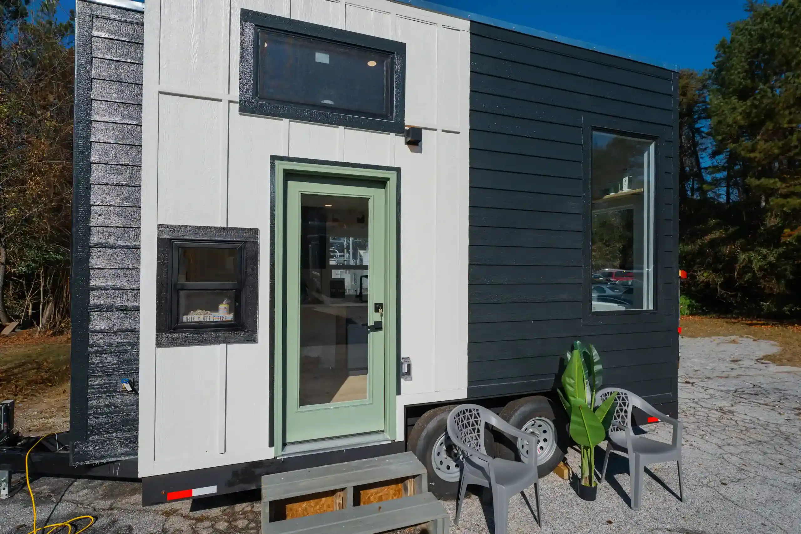 Sora on a dual-axle trailer under a blue sky: white board-and-batten, sage-green door, charcoal siding, wooden steps, chairs and plant on the lot