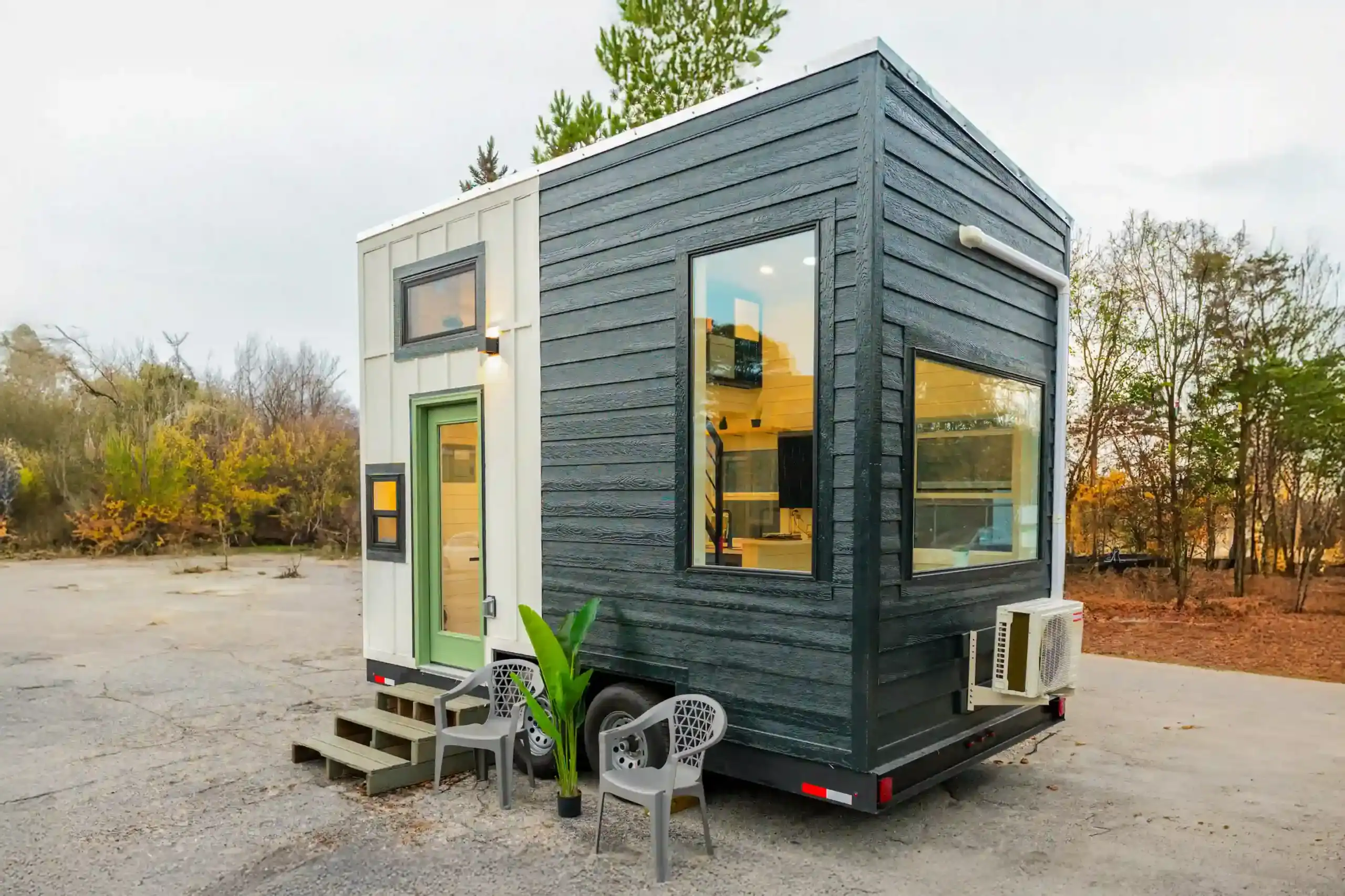 Three-quarter view of the Sora on a dual-axle trailer: sage-green door, large end picture window, white mini-split on the far wall, chairs and plant on gravel