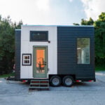 Two-tone 16-foot Sora tiny house on a black dual-axle trailer: white board-and-batten center, sage-green glass door, charcoal lap siding, black-framed windows, parked on asphalt under trees