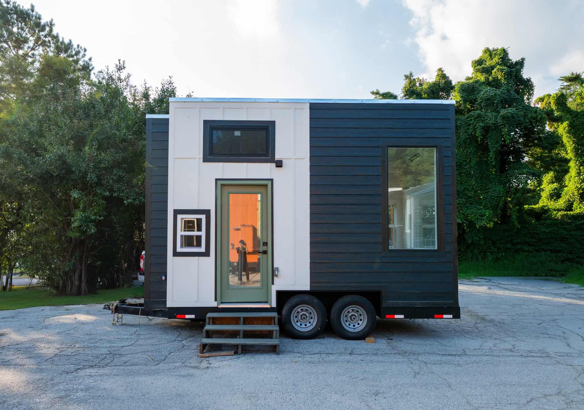 Two-tone 16-foot Sora tiny house on a black dual-axle trailer: white board-and-batten center, sage-green glass door, charcoal lap siding, black-framed windows, parked on asphalt under trees