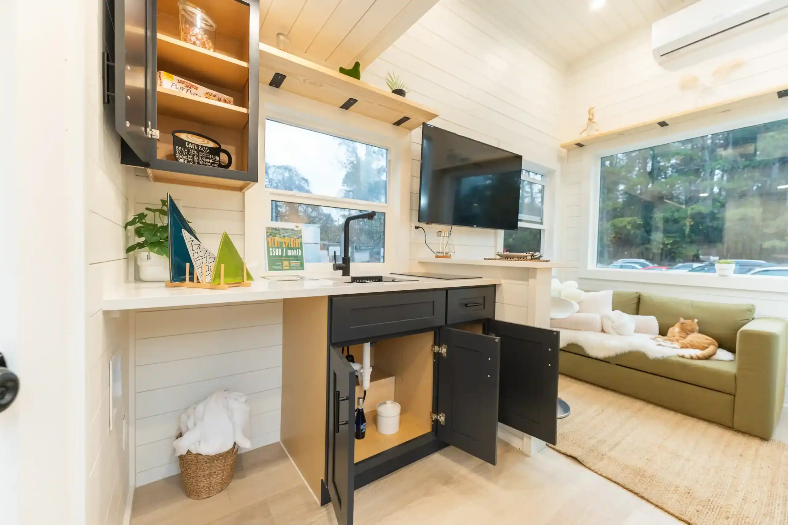 Sora kitchen: white counter, black faucet, two-burner cooktop, open dark base cabinets, olive sofa under the rear picture window