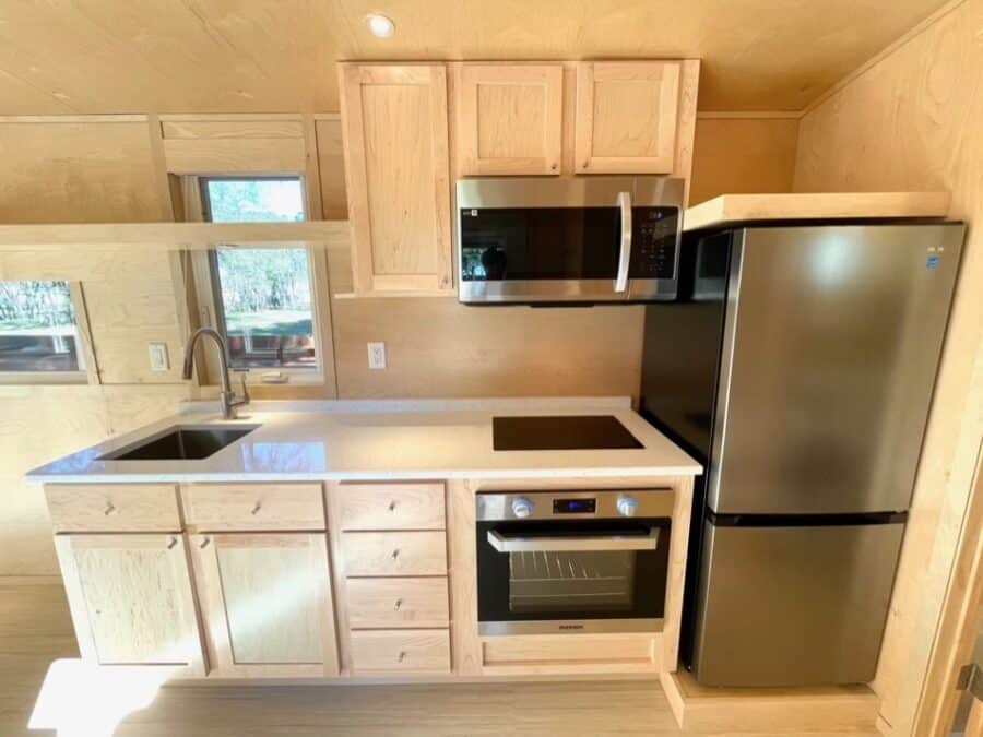 Compact tiny house kitchen featuring a refrigerator, oven, microwave, and wooden cabinets.