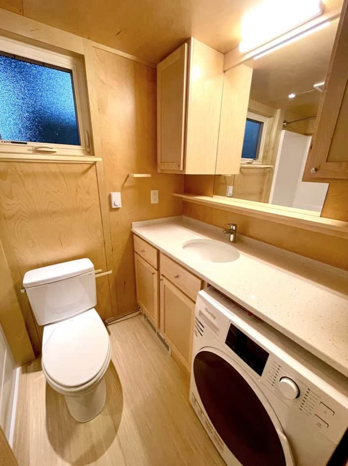 Compact tiny house bathroom featuring a washer, wooden cabinets, and a frosted window for natural li.