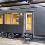 Black tiny house on wheels with large windows and a side door, inside visible through the windows.