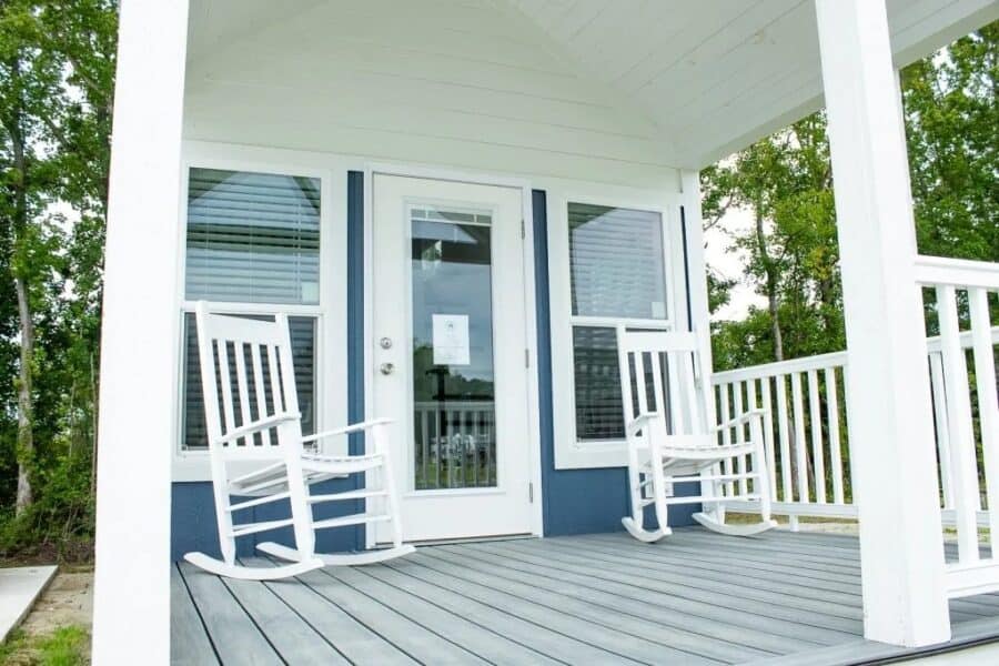 Covered porch of a blue and white park model with two white rockers and trees