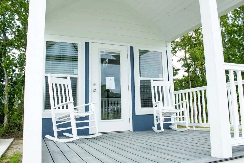 Covered porch of a blue and white park model with two white rockers and trees