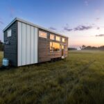 Tiny house on wheels with modern design in a grassy field during sunset.
