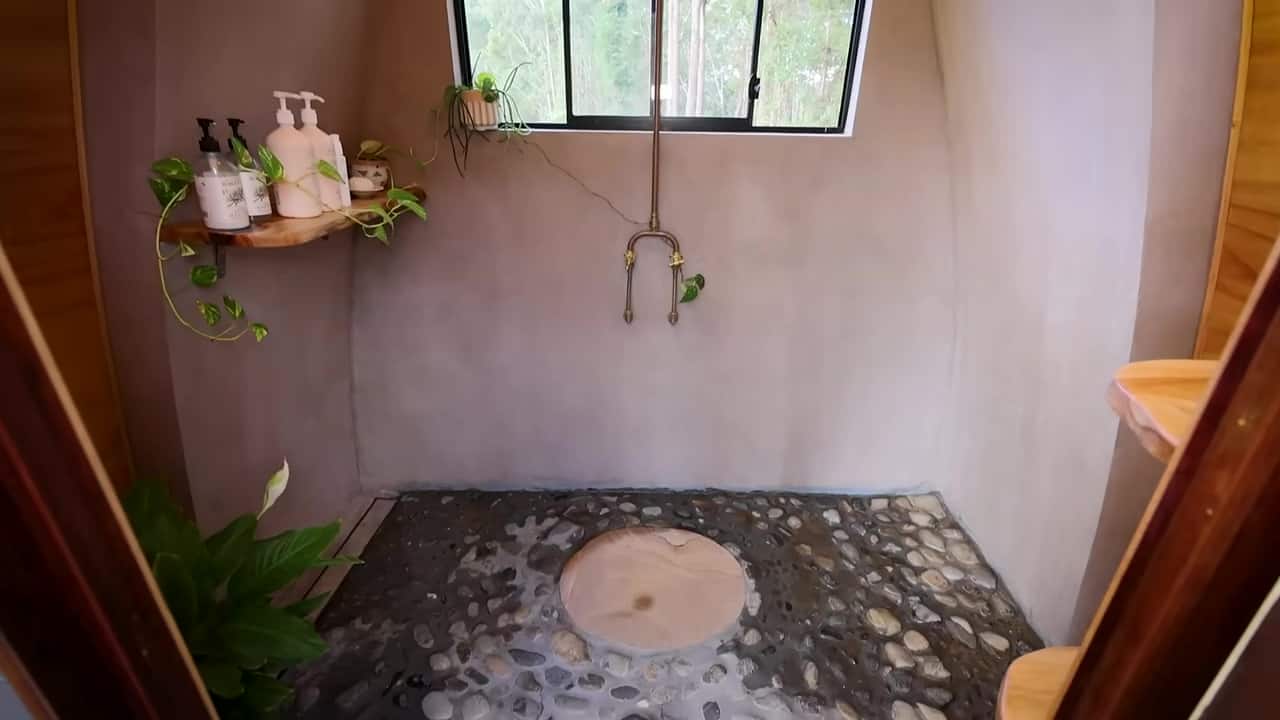 Curved plastered shower with a river-stone floor, sandstone pad, copper pipe, forest window, and plants