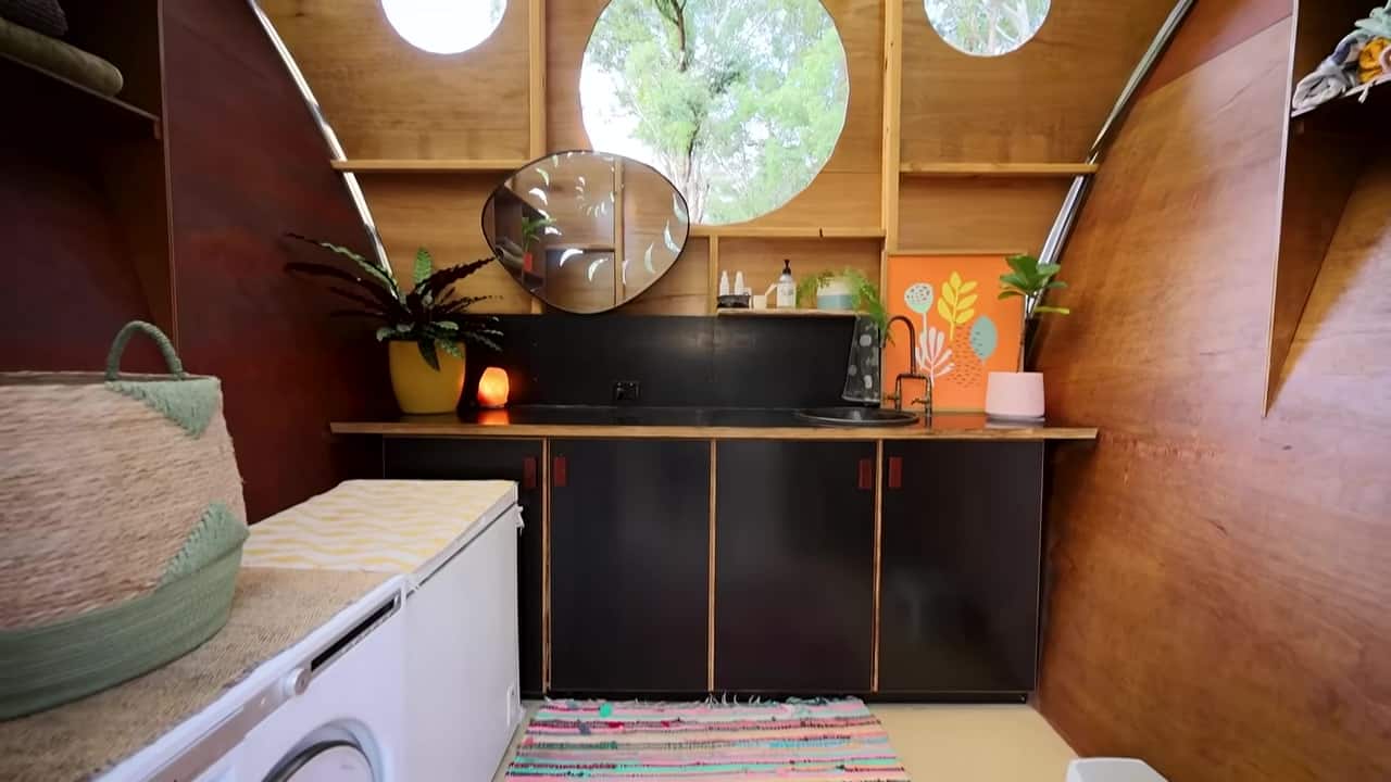 Curved plywood amenities interior with porthole windows, a black bench and round sink, a front-loader, and a striped rug