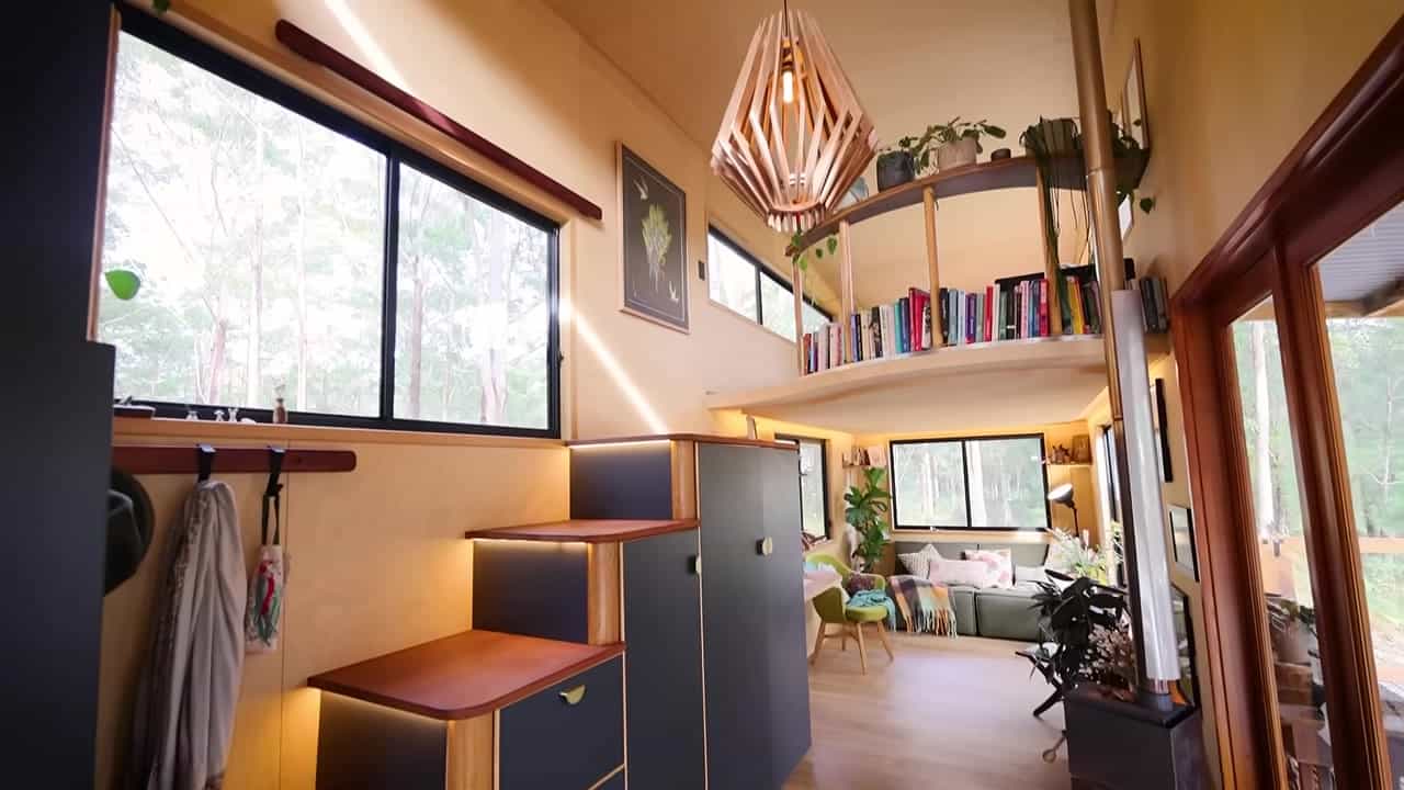 Open tiny-house interior with a sculptural storage stair, curved loft book rail, green sofa, wood-stove flue, and glass doors to a deck