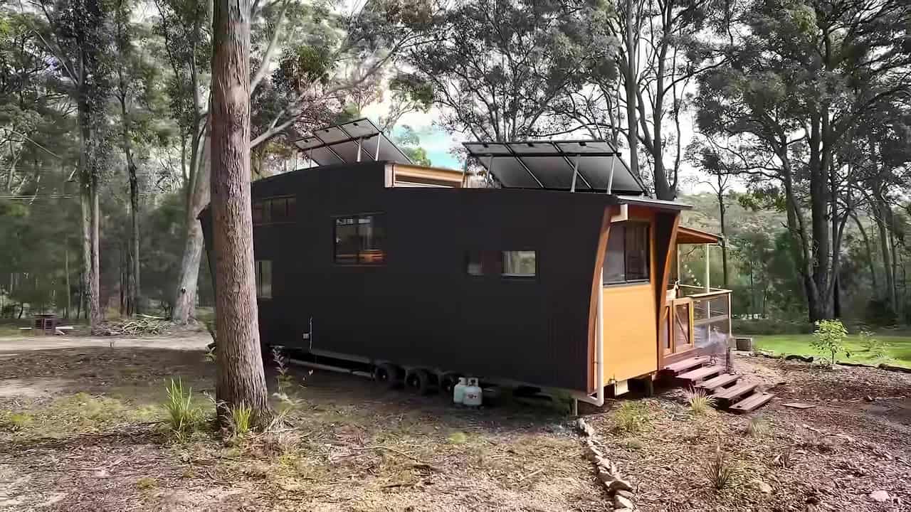 Side of a dark corrugated curved tiny house on a triple-axle trailer with wheels visible and solar on the roof