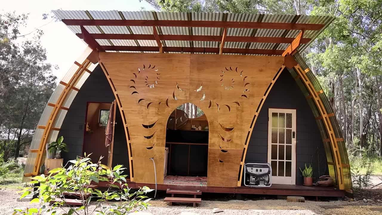 Curved amenities building with a plywood moon-cutout facade, arched doorway, laundry inside, and a timber deck in the bush