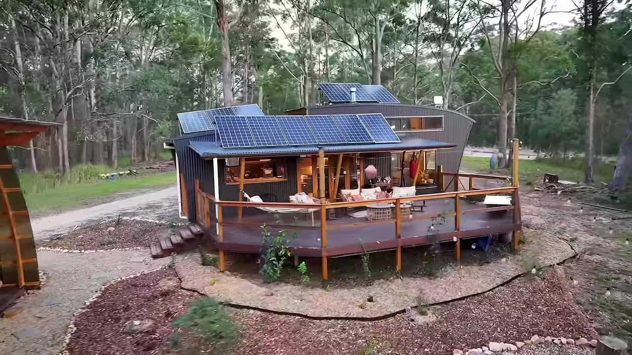 Aerial of a dark curved tiny house and oversized covered deck with solar panels in a eucalyptus clearing