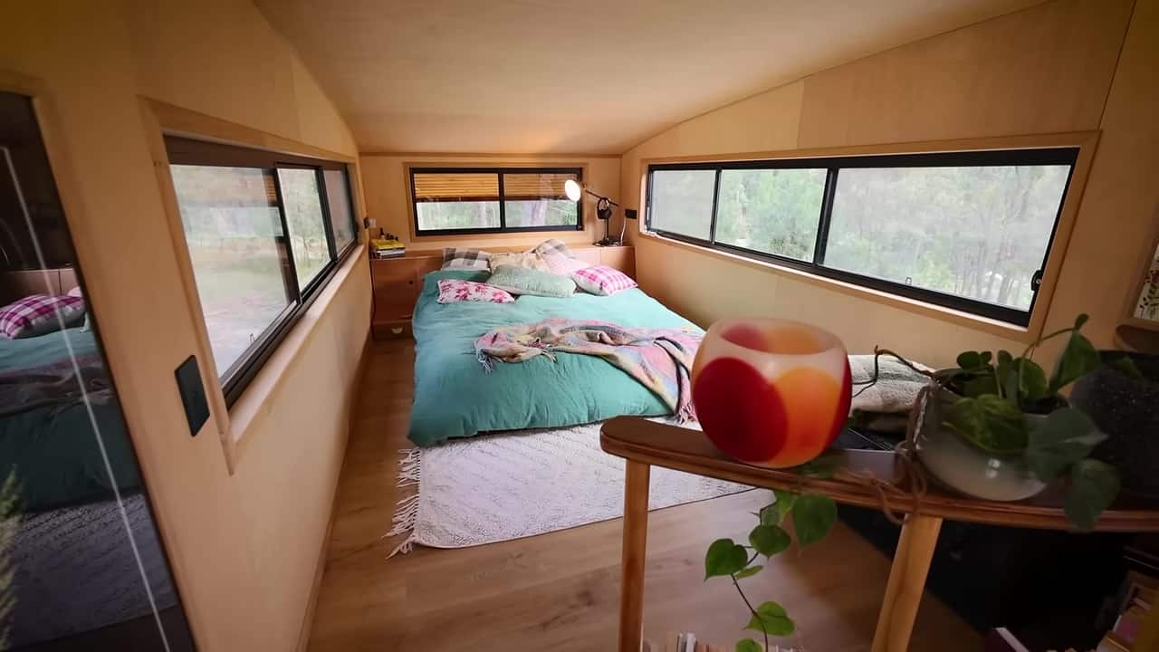 Standing-height tiny-house loft with a teal bed, timber headboard storage, and windows into the trees