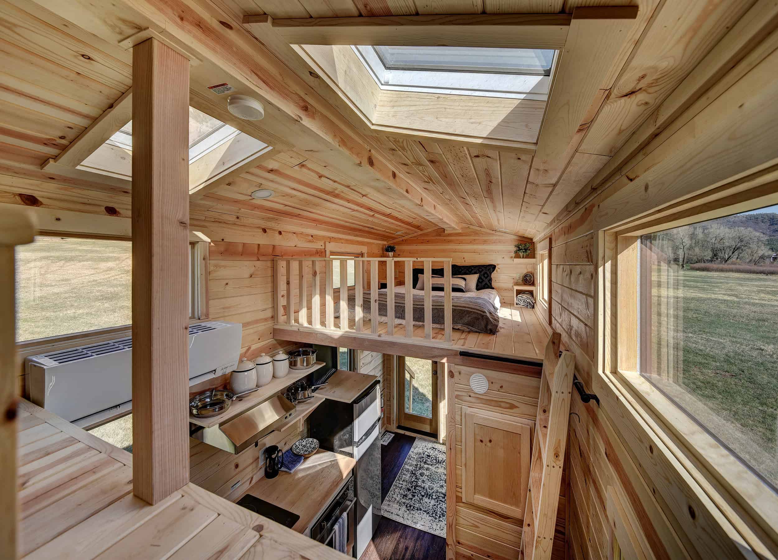 Tiny-house interior with light wood, a loft bed under a skylight, kitchen below, and field windows at Escalante Village in Durango