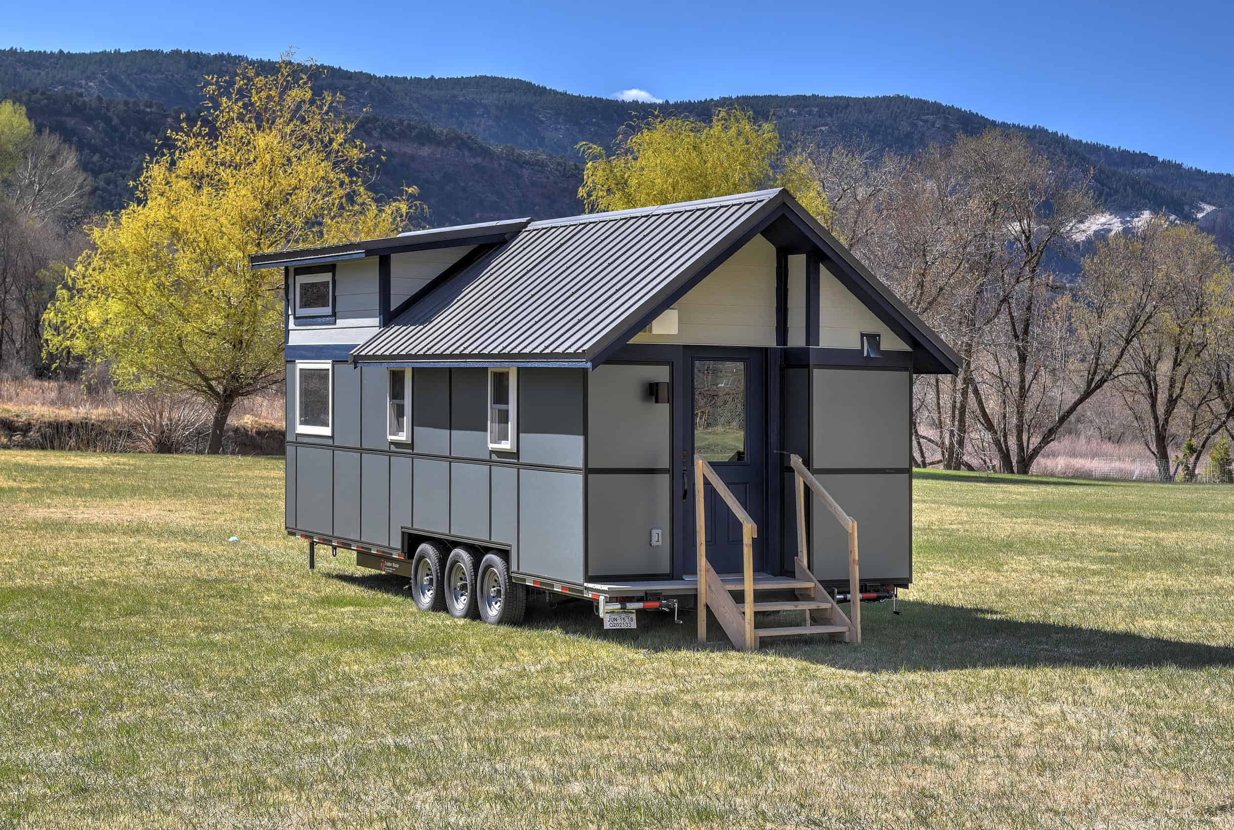 Solaire tiny house with dark panels, a cream gable, a blue door, and a triple-axle trailer on grass at Escalante Village in Durango