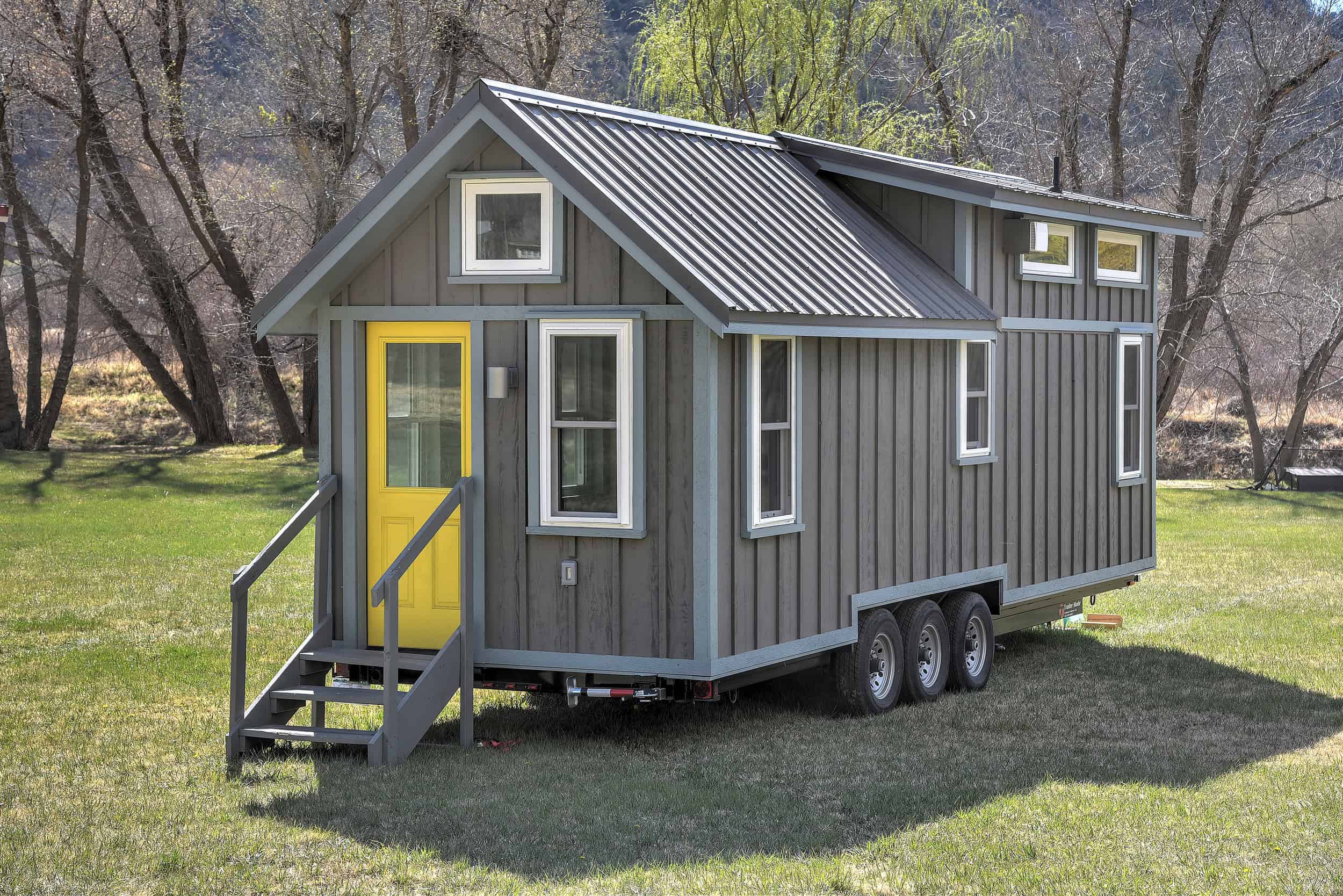 Sweetgrass tiny house with a yellow door, vertical siding, and a triple-axle trailer on grass at Escalante Village in Durango